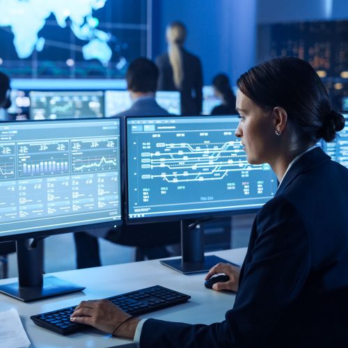 Confident Female Data Scientist Works on Personal Computer Wearing a Headset in Big Infrastructure Control and Monitoring Room. Woman Engineer in a Call Center Office Room with Colleagues.